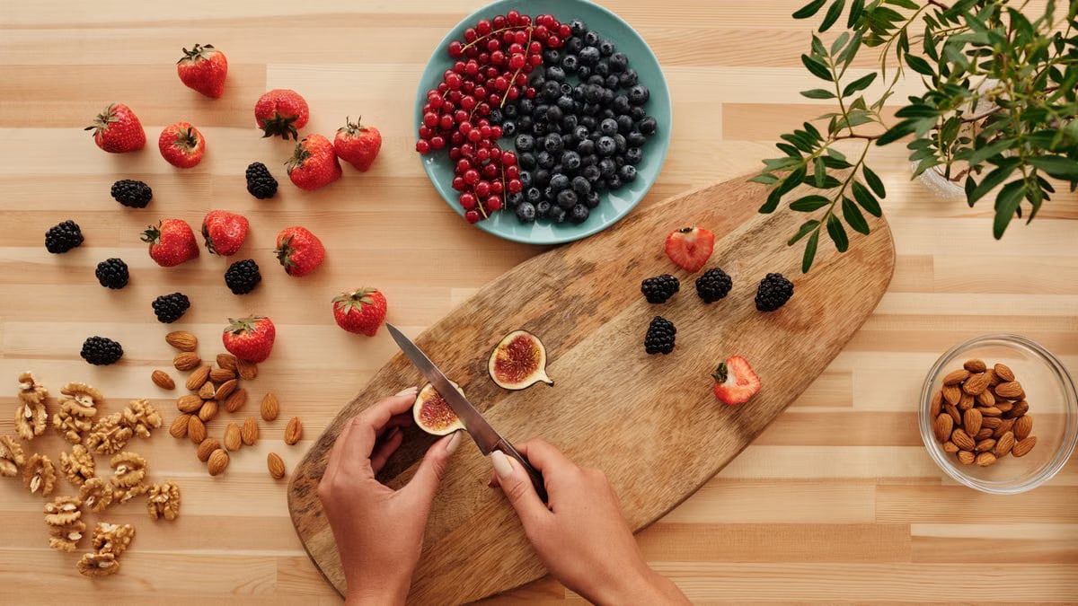 Overhead shot of fresh fruits, nuts, and hands preparing ingredients on a wooden board.